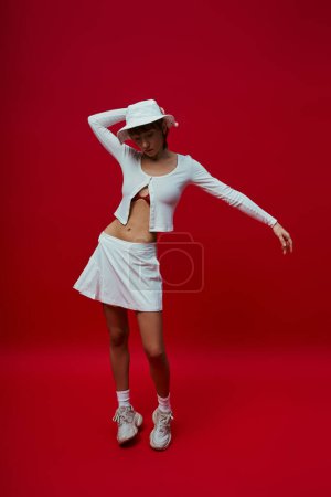 Foto de Mujer joven con estilo en traje blanco inteligente y sombrero contra fondo rojo vivo. - Imagen libre de derechos