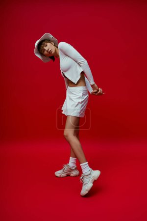 Elegant woman in white shirt and tennis skirt on red backdrop.