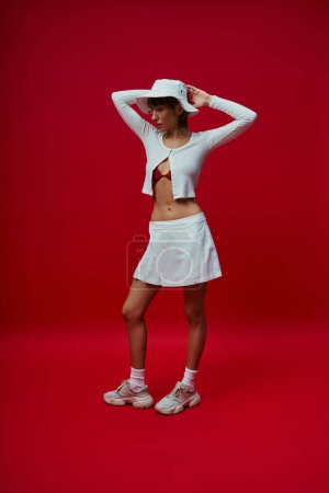 Foto de Una atractiva joven con una falda blanca y un sombrero se destaca sobre un vibrante telón de fondo rojo. - Imagen libre de derechos