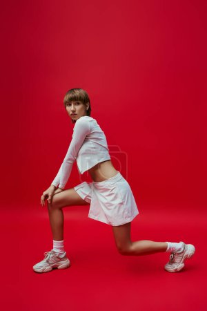Eine junge Frau in weißem Kleid und Tennisschuhen steht selbstbewusst vor einem leuchtend roten Hintergrund..