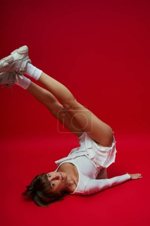 Young woman in white sportswear performs yoga on vibrant red background.