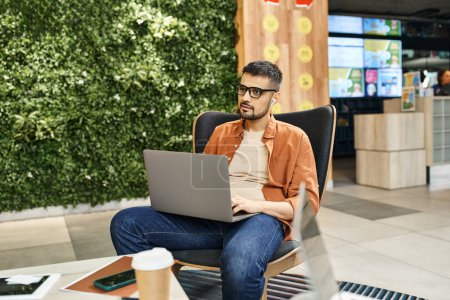 Un hombre en una silla, absorto en su portátil, rodeado por el zumbido de un espacio de coworking donde los miembros del equipo de startups colaboran.