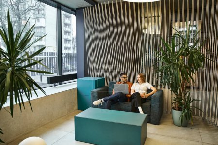 Two professionals from a startup team sit on a trendy couch in a contemporary office space, taking a break from work.