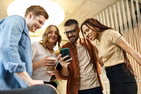 Un grupo diverso de colegas en un espacio de coworking moderno se reúnen alrededor de un teléfono celular, absortos en lo que ven.