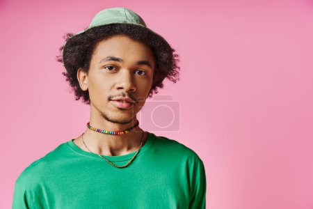 Cheerful African American man with curly hair dons a green shirt and matching hat against a vibrant pink backdrop.