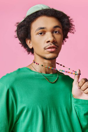 A young African American man with curly hair wearing a green shirt and a beaded necklace on a pink background.