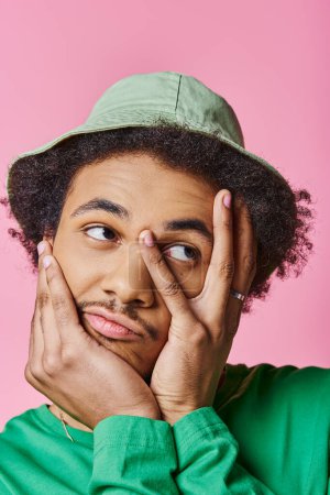 Photo for A pensive African American man in casual wear, with curly hair, wearing a green shirt and hat, on a pink background. - Royalty Free Image