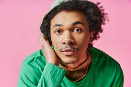 young African American man with curly hair wearing a green shirt and hat, on a pink background.