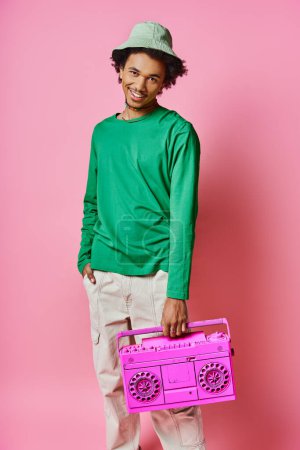 Curly African American man in a green shirt joyfully holding a pink radio on a pink background.