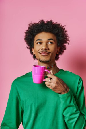 A cheerful, young African American man in casual wear holds a cup in front of his face against a pink background.