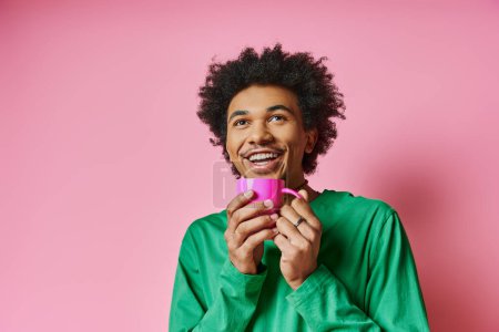Ein fröhlicher junger afroamerikanischer Mann in Freizeitkleidung hält eine rosa Tasse vor einem leuchtend rosa Hintergrund.
