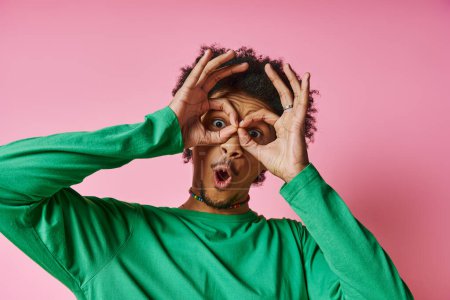 A young African American man in casual wear on a pink background, making a playful and expressive face using his hands.