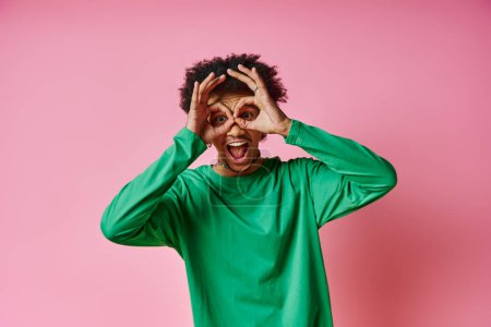 Un hombre afroamericano alegre con el pelo rizado en una camisa verde, sosteniendo alegremente sus manos hacia arriba a su cara sobre un fondo rosado.