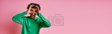 Cheerful young African American man in green shirt covering his eyes, expressing emotions, on a pink background.