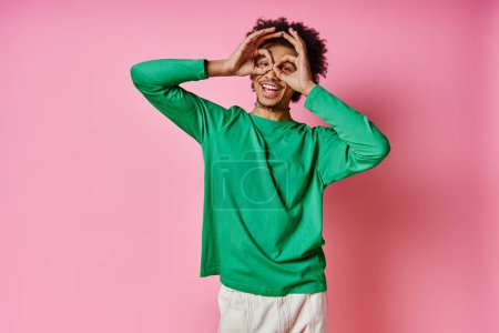 Fröhlicher junger afroamerikanischer Mann in grünem Hemd, der seine Augen verhüllt und fröhliche Emotionen auf rosa Hintergrund ausdrückt.