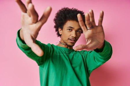 A cheerful young African American man with curly hair in a green shirt raising his hands in joy on a pink background.