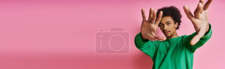 Cheerful curly African American man in casual green shirt with hands raised, expressing positivity on a pink background.