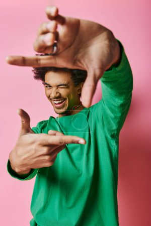 A cheerful African American man in a green shirt makes a gesture against a pink background.