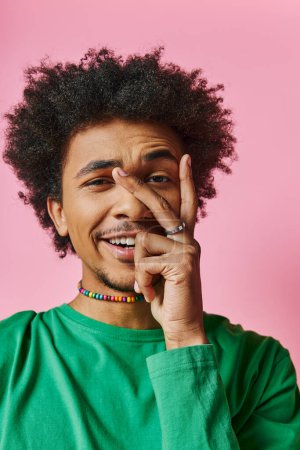 Foto de Alegre hombre afroamericano con el pelo rizado en una camisa verde, sonriendo y tocando su cara sobre un fondo rosa. - Imagen libre de derechos