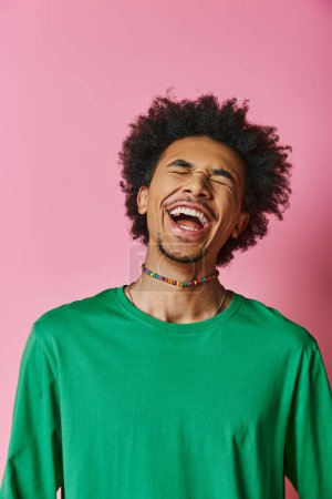A cheerful young African American man with a curly afro laughs while wearing a green shirt on a pink background.
