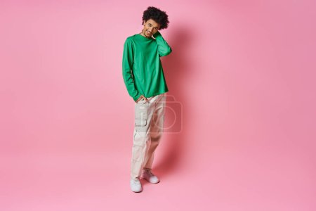 A cheerful young African American man in a green sweater stands against a pink background, exuding emotion.