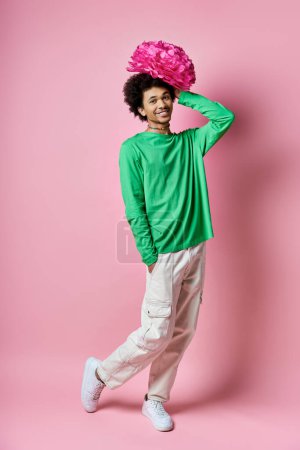 A cheerful young African American man in a green shirt and white pants, with a pink flower on his head, against a pink background.