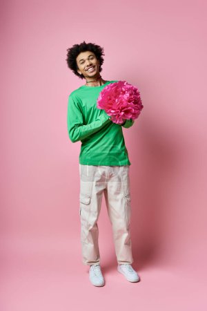 Cheerful young African American man in green shirt holding a delicate pink flower against a soft pink background.