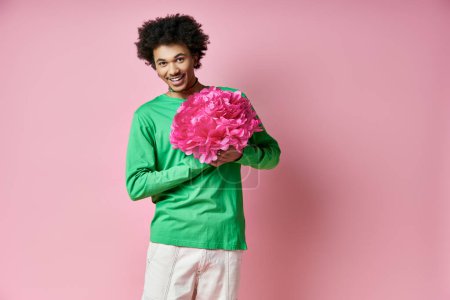 Foto de Hombre afroamericano alegre con camisa verde sosteniendo una flor rosa, expresando emociones sobre un fondo rosa. - Imagen libre de derechos