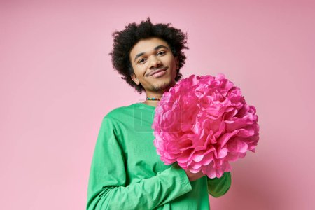 A cheerful, curly-haired African American man in casual wear holds a large pink flower against a pink background.