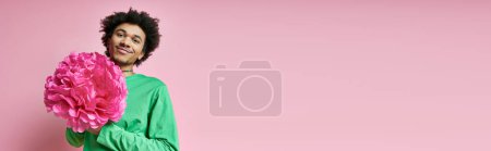 Téléchargez les photos : Joyeux homme afro-américain aux cheveux bouclés, en chemise verte, tenant une délicate fleur rose sur un fond rose vibrant. - en image libre de droit