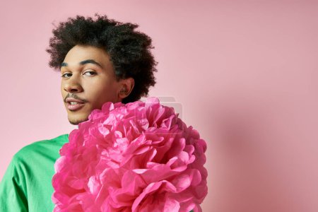 A cheerful young African American man wearing casual attire holds a large pink flower in front of his face, showcasing emotion and elegance.
