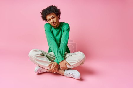 A cheerful young African American man with curly hair sits cross-legged on the ground, deep in thought.