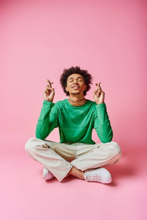A cheerful young African American man in a green shirt strikes a yoga pose with focus and grace on a pink background.