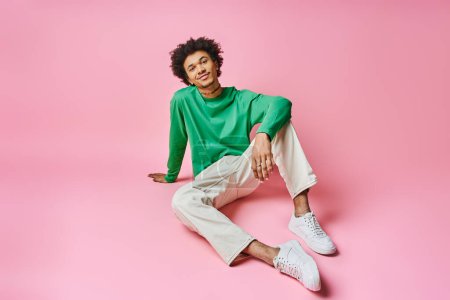 A cheerful African American man with curly hair sitting on a pink surface, wearing a green shirt and white pants.