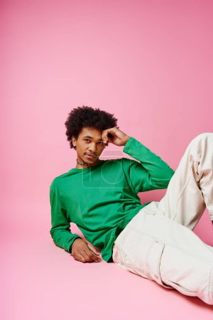 Cheerful African American man with curly hair in casual green shirt and white pants, expressing emotions on pink background.