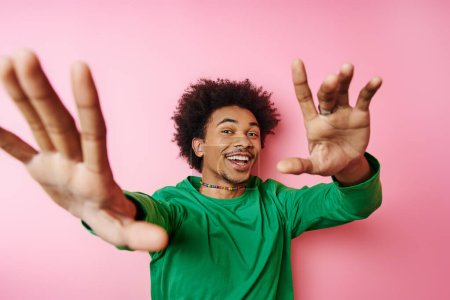 Young, curly-haired man in casual attire, raising his hands in excitement on a pink background.