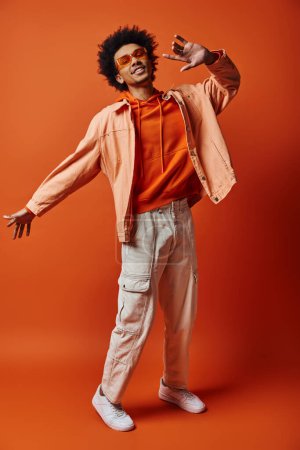 Foto de Un joven afroamericano de moda con el pelo rizado posa con confianza en camisa y pantalones blancos sobre un fondo naranja vibrante. - Imagen libre de derechos