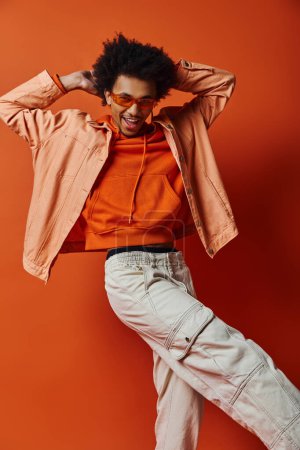 Foto de Stylish young African American man in orange shirt and white pants, wearing sunglasses, exuding confidence on orange background. - Imagen libre de derechos