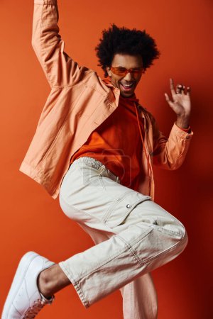 Curly African American man in trendy outfit and sunglasses, jumping energetically in the air against orange background.