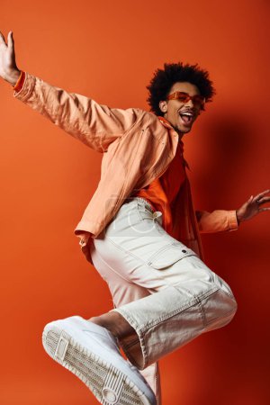 Foto de Un joven afroamericano enérgico salta en el aire con los brazos extendidos, exhalando alegría y libertad contra un fondo naranja. - Imagen libre de derechos