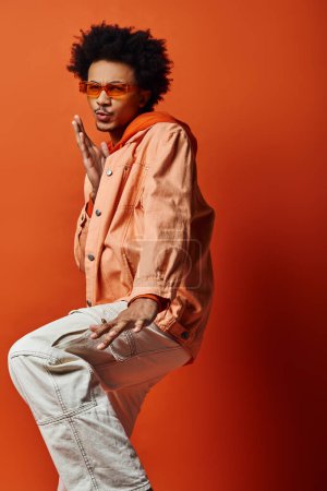 Curly African American man standing gracefully on one leg, exuding confidence and style, wearing trendy attire, on orange background.