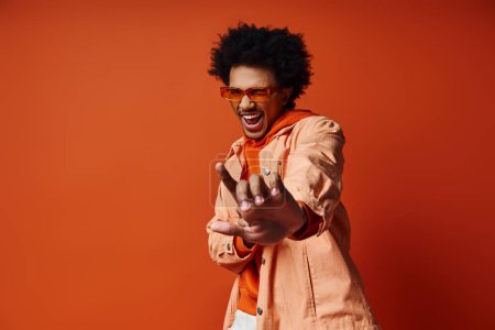Téléchargez les photos : Un homme afro-américain élégant aux cheveux bouclés portant une chemise orange et un pantalon blanc, mettant en valeur la mode à la mode sur un fond orange. - en image libre de droit