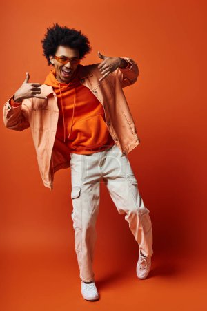 Foto de Elegante joven rizado hombre en camisa naranja de moda y pantalones blancos posando emocionalmente sobre un fondo vibrante. - Imagen libre de derechos