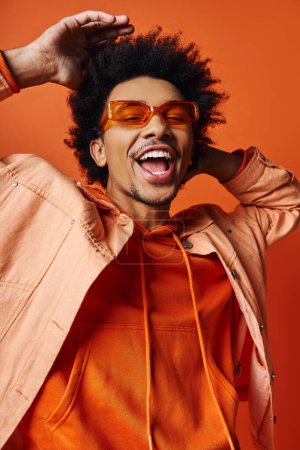 Foto de Un joven afroamericano elegante con el pelo rizado con una sudadera con capucha naranja y gafas de sol sobre un fondo naranja vibrante, mostrando emociones. - Imagen libre de derechos