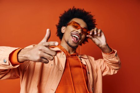 Ein stilvoller junger afroamerikanischer Mann in orangefarbenem Hemd und Sonnenbrille, der auf orangefarbenem Hintergrund ein albernes Gesicht macht.