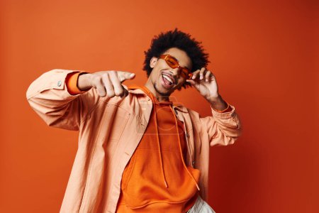 A stylish young African American man with curly hair wearing an orange shirt and sunglasses, posing energetically against an orange background.