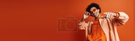 Photo for A stylish young African American man in an orange shirt and sunglasses holding his hand to his face on a vibrant orange background. - Royalty Free Image
