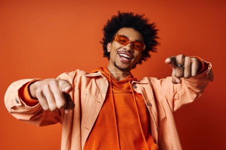 Stylish African American man in orange shirt and sunglasses pointing towards camera on vibrant orange background.