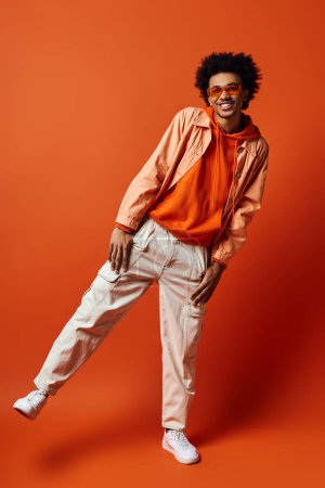 Un joven y elegante hombre afroamericano con el pelo rizado con una camisa naranja y pantalones caqui, gafas de sol deportivas sobre un fondo naranja.