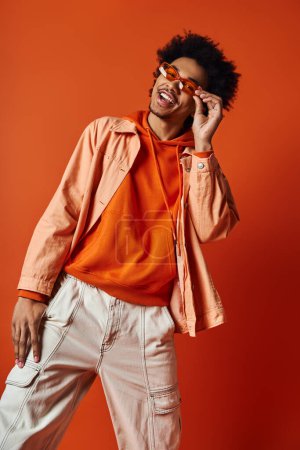 Hombre afroamericano con pelo afro rizado y gafas de sol posando expresivamente en una camisa naranja.